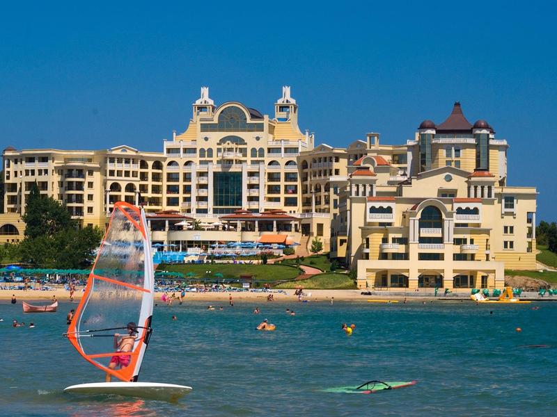 Hotel di lusso sul mare con spiaggia e surfista in primo piano sotto un cielo limpido.