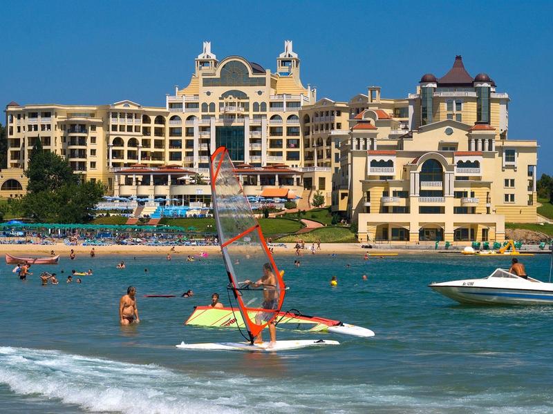 Hotel di lusso sulla spiaggia con barche a vela e nuotatori nel mare sotto un cielo limpido.
