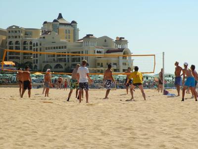 Meerdere mensen spelen beachvolleybal op een zandstrand met gebouwen op de achtergrond.