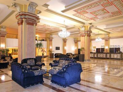 Ruime hotellobby met marmeren vloer, zuilen en blauwe zitgroepen onder decoratief plafond.