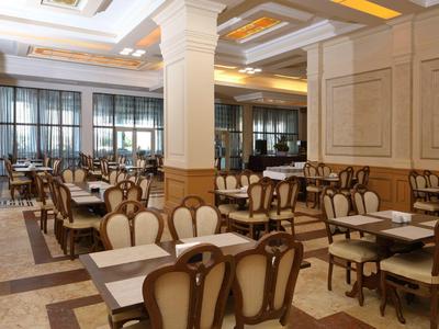 Elegant hotelrestaurant met gestoffeerde stoelen, witte tafels en grote ramen onder decoratieve plafondverlichting.