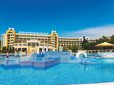Groot meerlaagse hotel met blauw zwembad en waterfonteinen onder een heldere hemel.