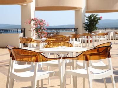 Terras met witte tafels en bruine stoelen met uitzicht op een zwembad en de zee.