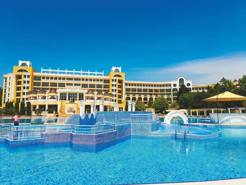 Groot meerlaagse hotel met blauw zwembad en waterfonteinen onder een heldere hemel.