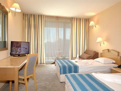 Lichte hotelkamer met twee bedden, bureau, stoel en groot raam met gordijnen.