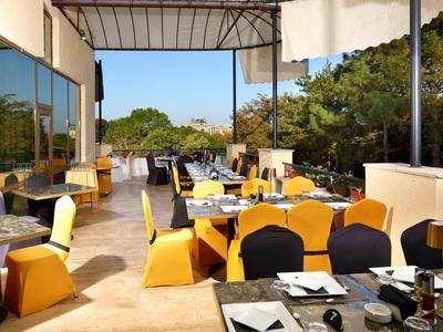 Open terras van een restaurant met tafels en stoelen, omgeven door bomen en blauwe lucht.