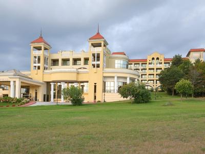 Velká žlutá hotelová budova s věžemi před zalesněným kopcem a travnatou plochou.