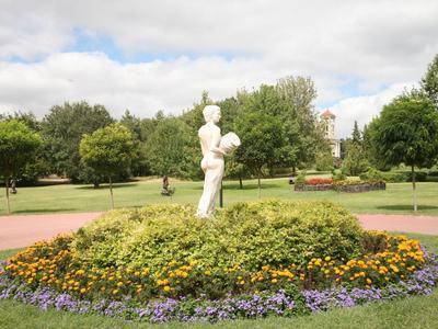 Witte standbeeld van een figuur in een parkachtige tuin met bloembedden en bomen op de achtergrond