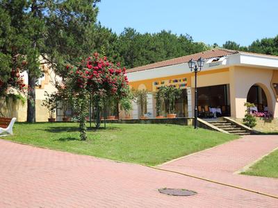 Malá žlutá budova s trávníkem, stromy a červenými květy v hotelovém areálu.