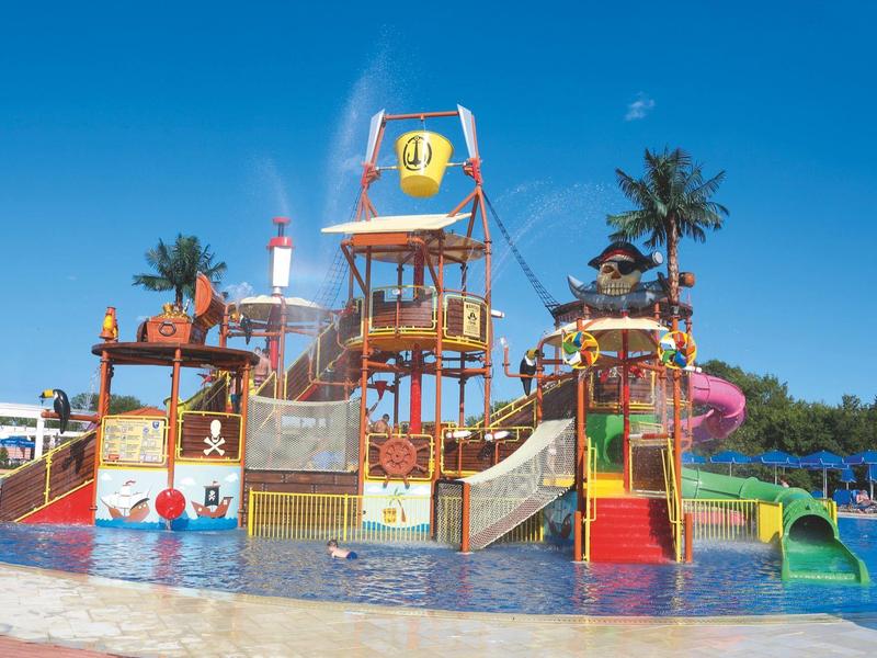 Kleurrijke waterspeeltuin met glijbanen, sproeiers en palmbomen in een ondiep zwembad onder een blauwe lucht
