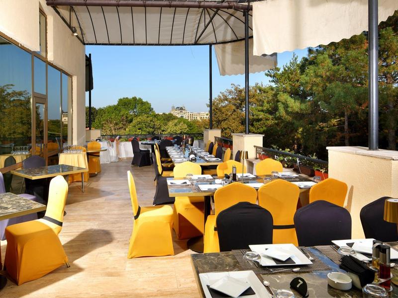 Open terras van een restaurant met tafels en stoelen, omgeven door bomen en blauwe lucht.