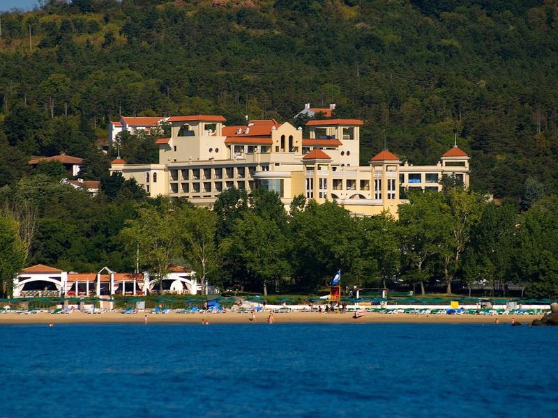 Groot hotel aan het water met bomen en heuvels op de achtergrond bij heldere lucht.