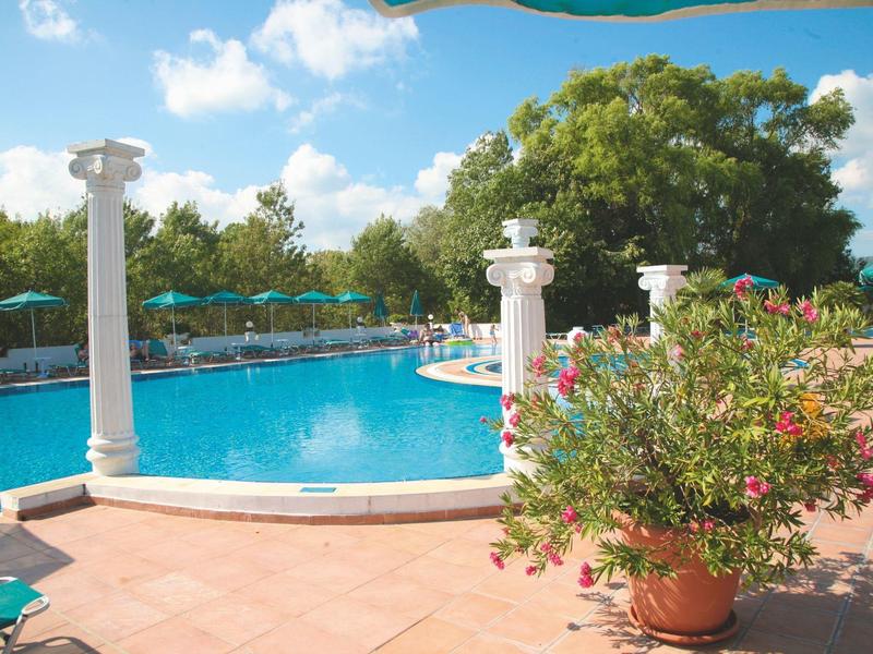 Zwembad met blauwe parasols en witte zuilen, omgeven door bomen en een heldere blauwe lucht.