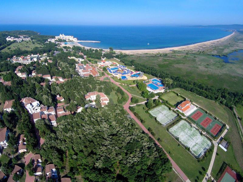 Luchtfoto van een strandresort met tennisbanen, zwembaden en bosrijke gebieden bij de zee.