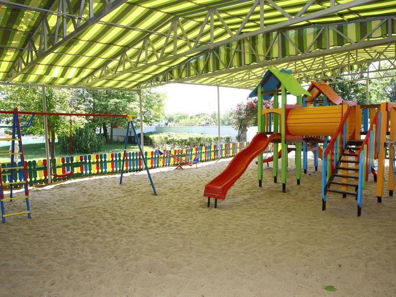 Overdekte speeltuin met zandgrond, kleurrijke glijbaan, schommels en klimrek.