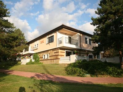 Modernes zweistöckiges Haus mit Balkon, umgeben von Bäumen und grüner Wiese bei sonnigem Himmel.