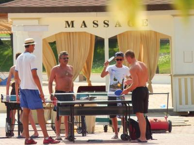 Fünf Männer spielen Tischfußball vor einem gelben Massagezelt im Freien bei Sonnenschein.