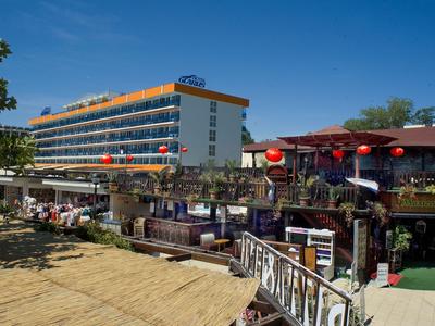 Hotelová budova s balkony vedle restaurace a písečné pláže pod jasně modrou oblohou