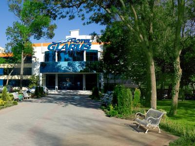 Moderní hotelová budova obklopená stromy a zelenými trávníky, s lavičkami podél cesty.