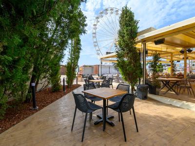 Venkovní prostor kavárny s stolky a židlemi, v pozadí ruské kolo a stromy.