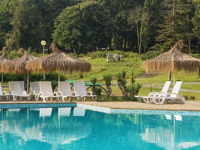 Großer blauer Pool mit weißen Liegestühlen und Strohdach-Sonnenschirmen, umgeben von Bäumen.