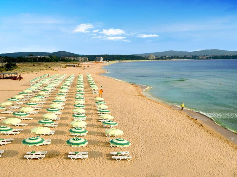 Wide sandy beach with sun loungers and umbrellas by calm sea under blue sky.