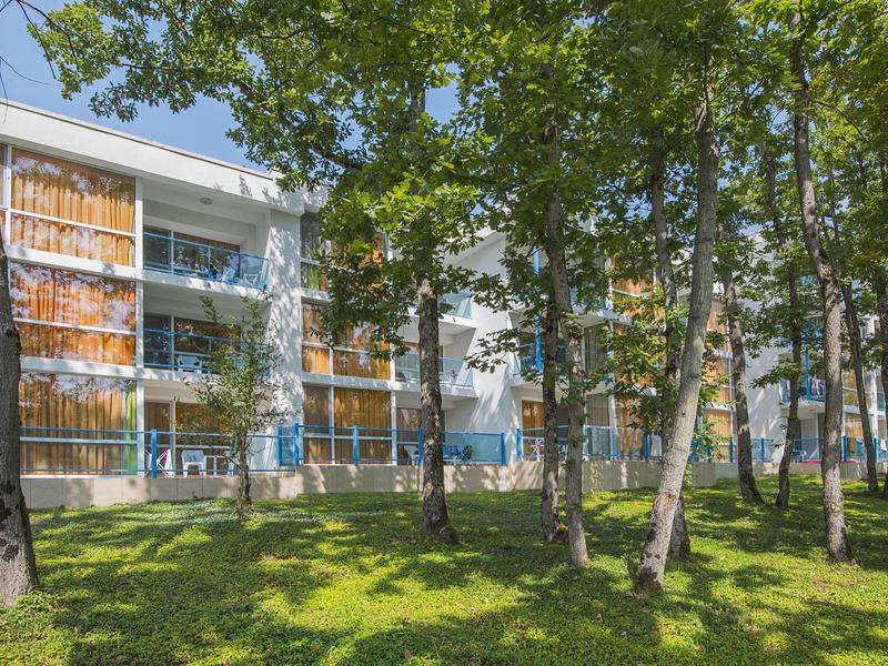 Multi-story residential building with balconies behind a park with trees on a lawn