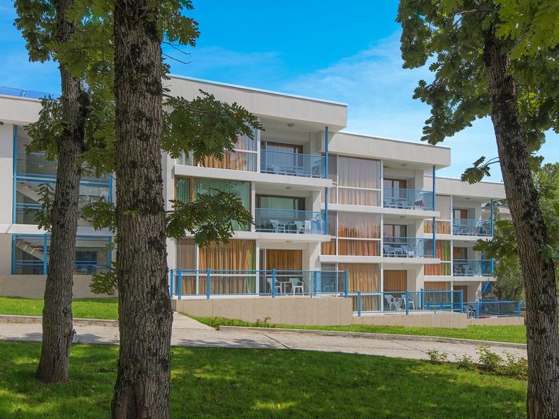 Modern hotel building with balconies and trees in the foreground under a blue sky.