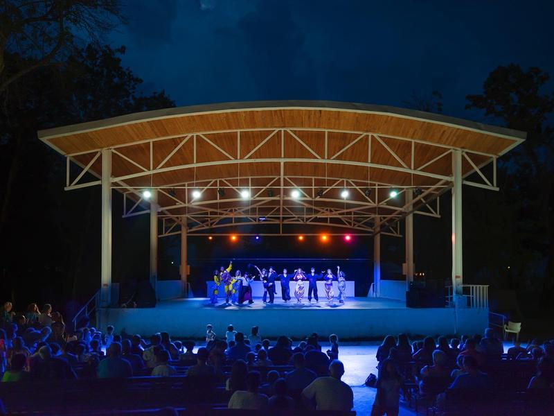 Illuminated stage with band at night, outdoor audience seated in front.