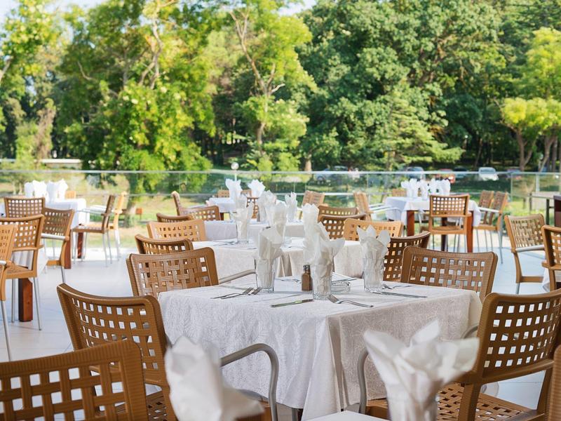 Restaurant terrace with white tablecloths and chairs overlooking green trees and water.