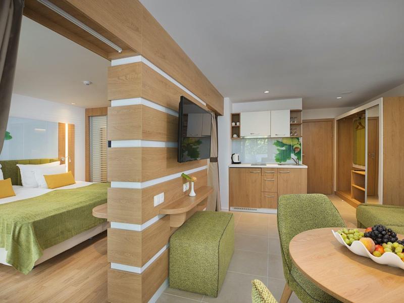 Modern hotel room with green decor, bedroom, living area, and kitchenette.