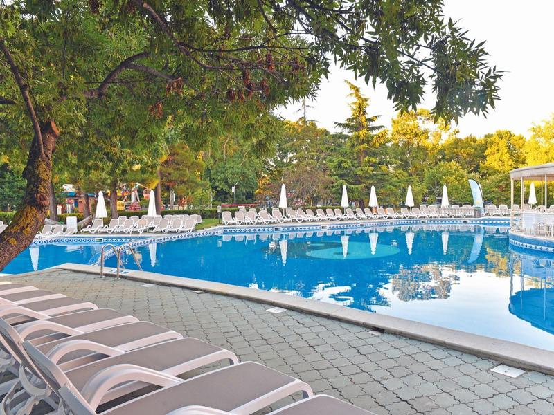 Large hotel pool with sun loungers and umbrellas in a green setting.