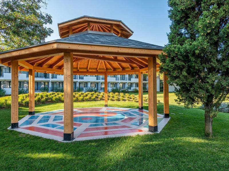 Open wooden gazebo on green lawn beside a tree in a garden.