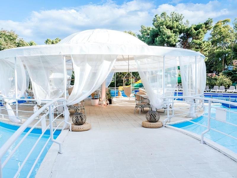 White pavilion with sheer curtains beside a blue hotel pool in a sunny setting.
