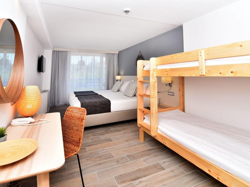 Bright hotel room with double bed, bunk bed, desk, and chair in warm wood tones.