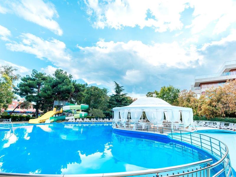 Large pool with water slide and white tent in a hotel garden under cloudy sky.