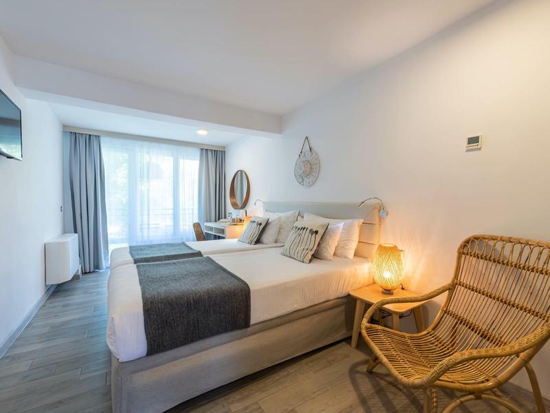 Bright hotel room with double bed, wooden floor, wicker chair, and balcony with curtains.