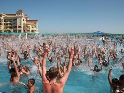 Viele Menschen genießen das Schwimmbad mit erhobenen Armen vor einem Hotelgebäude bei klarem Himmel.