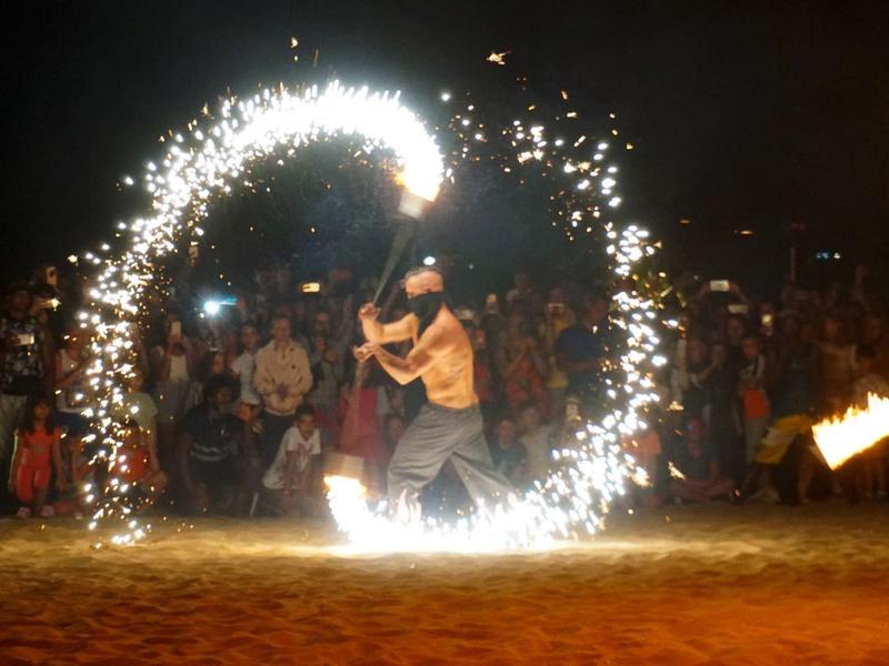 Spettacolo di fuoco sulla spiaggia di notte con scintille e pubblico sullo sfondo