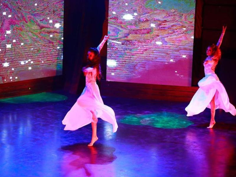 Due ballerine in abiti bianchi si esibiscono su un palco con effetti luminosi colorati.