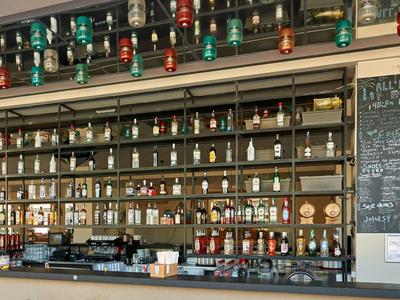 Bancone di bar con numerose bottiglie di bevande diverse e una lavagna con offerte.