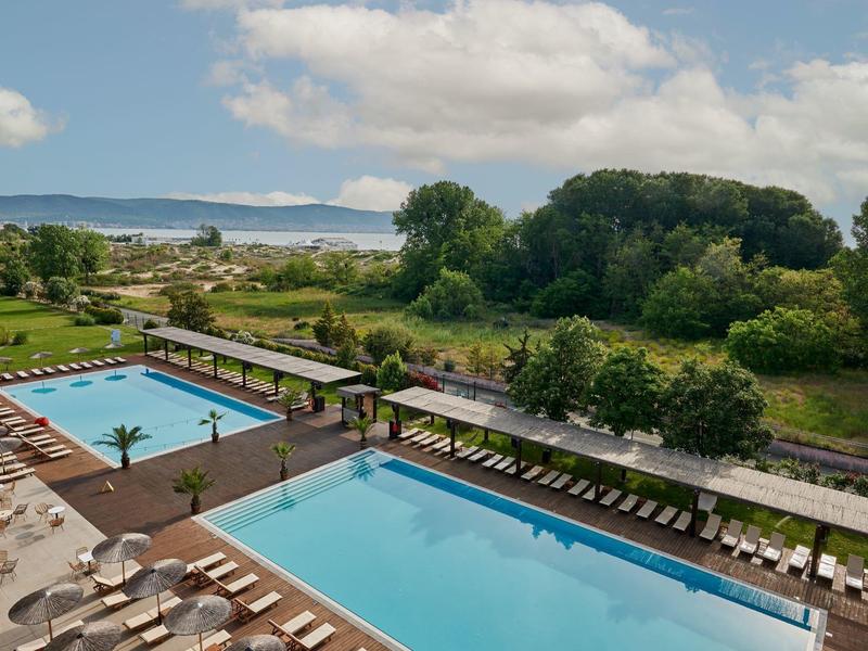 Buitenzwembaden met ligstoelen bij een hotel naast groen landschap en uitzicht op afstand.