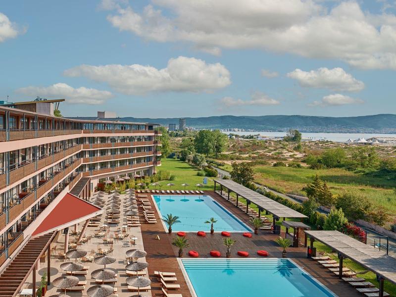 Hotel met twee buitenzwembaden, ligstoelen en uitzicht op groen en de zee.