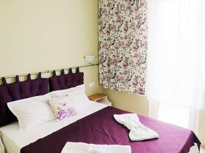 Schlafzimmer mit lila Bettdecke, weißen Kissen, floralem Vorhang und natürlichem Tageslicht.