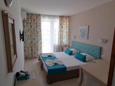 Hell eingerichtetes Schlafzimmer mit Doppelbett, blauen Kissen und Vorhängen, Fernseher und kleinem Kühlschrank.