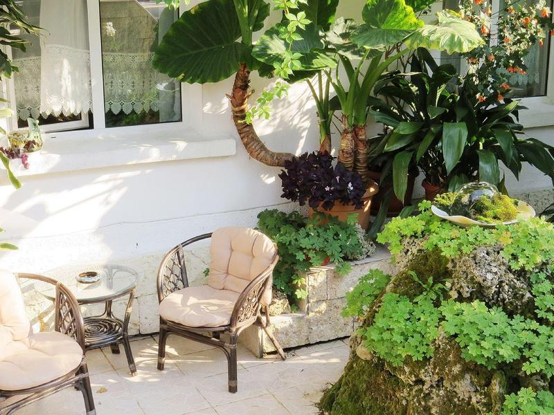 Kleiner Garten mit zwei Stühlen, Glastisch, großen grünen Pflanzen und weißen Hauswänden.