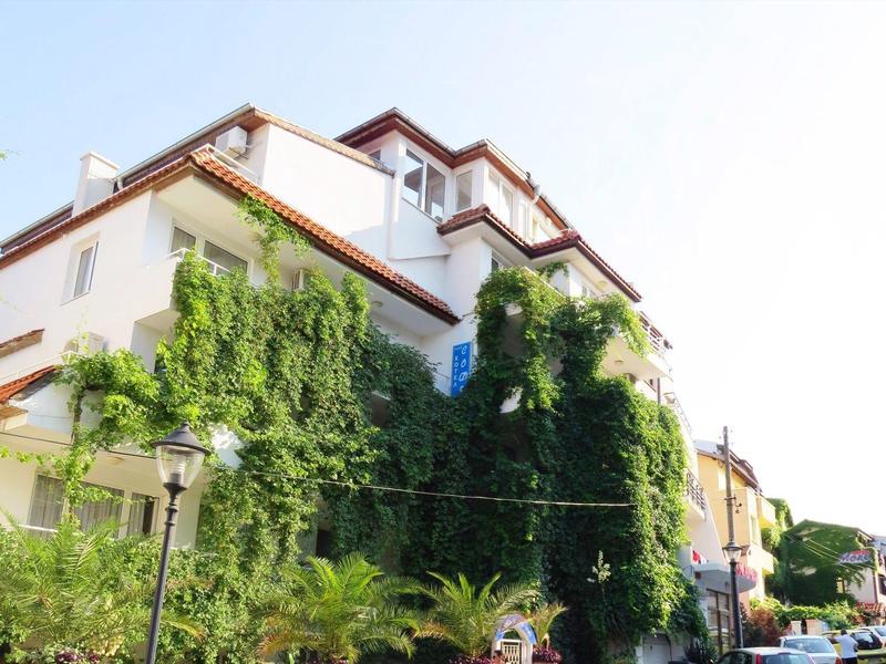 Mehrstöckiges weißes Haus mit vielen grünen Kletterpflanzen an der Fassade bei Sonnenschein.