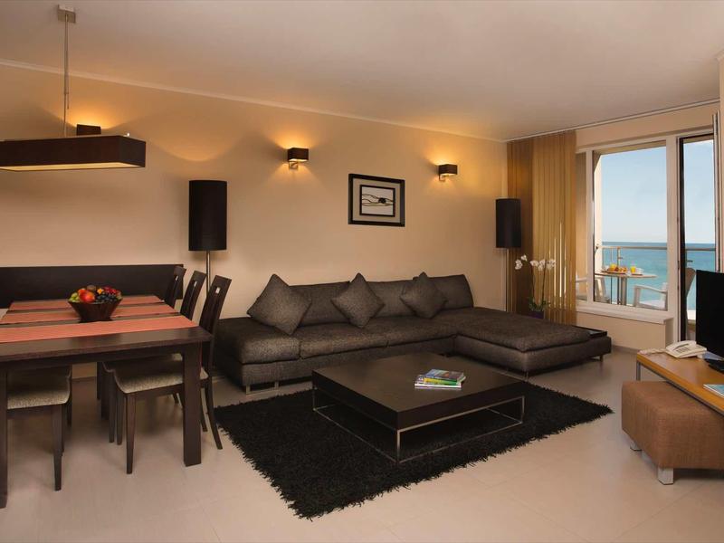 Helles Wohnzimmer mit großer Eckcouch, Holz-Essgruppe, TV und Meerblick durch Fenster.