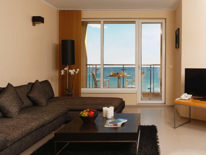 Helles Wohnzimmer mit grauem Sofa, schwarzem Teppich, Holztisch und großer Fensterfront zum Meer.