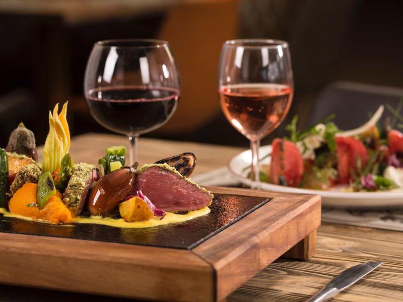 Serviertes Gourmet-Gericht mit Fleisch, Gemüse und zwei Weingläsern auf Holztisch.